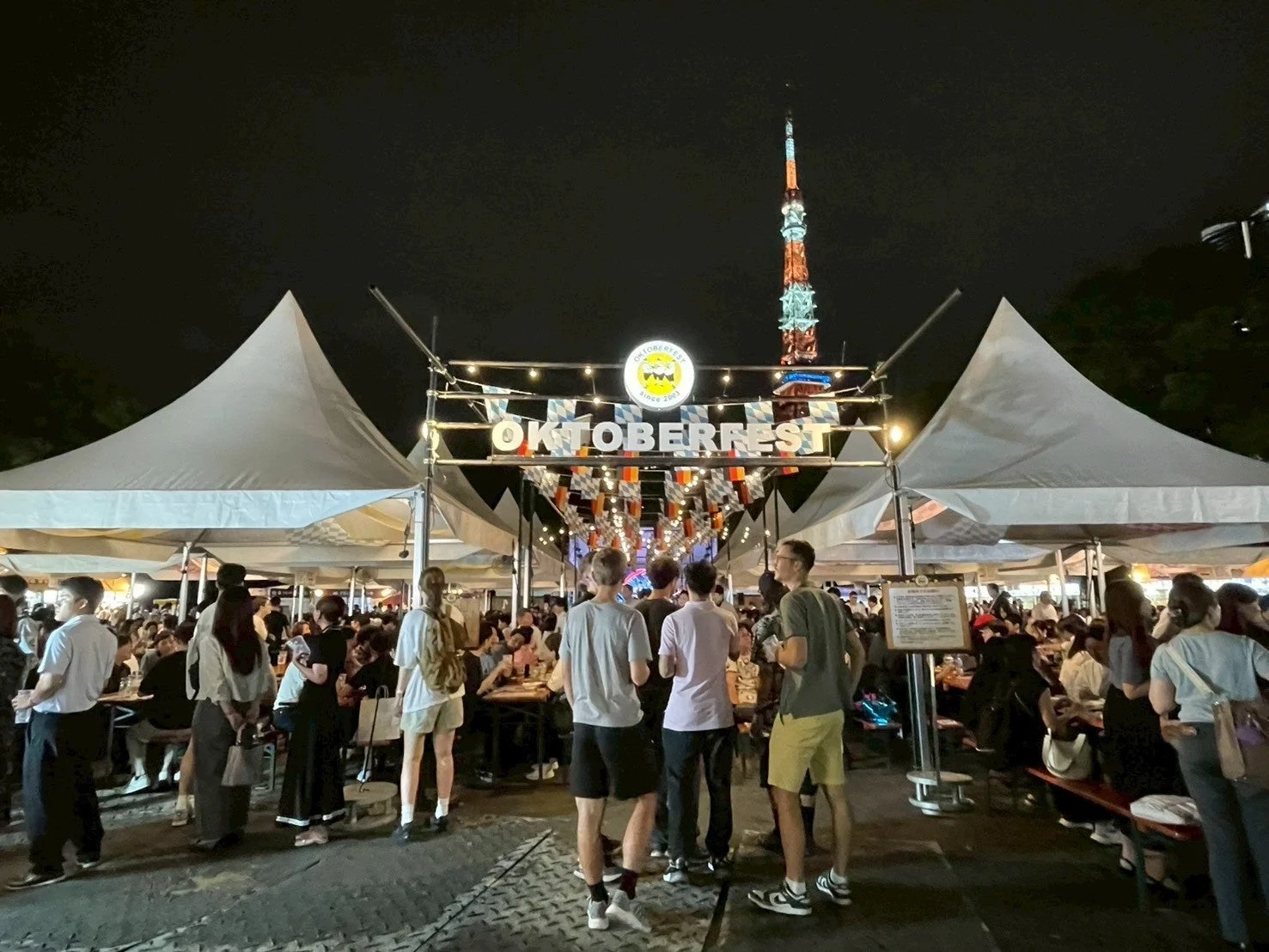 夜の屋外イベント会場の様子。多くの人々がオクトーバーフェストのゲートやテントの下に集まり、賑わっている。背景にはライトアップされた東京タワーが見える。