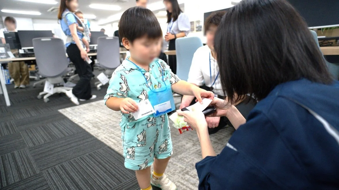 オフィス内で、子供が大人と交流している様子です。子供は社員証のようなカードを持っており、大人が何か手渡しています。
