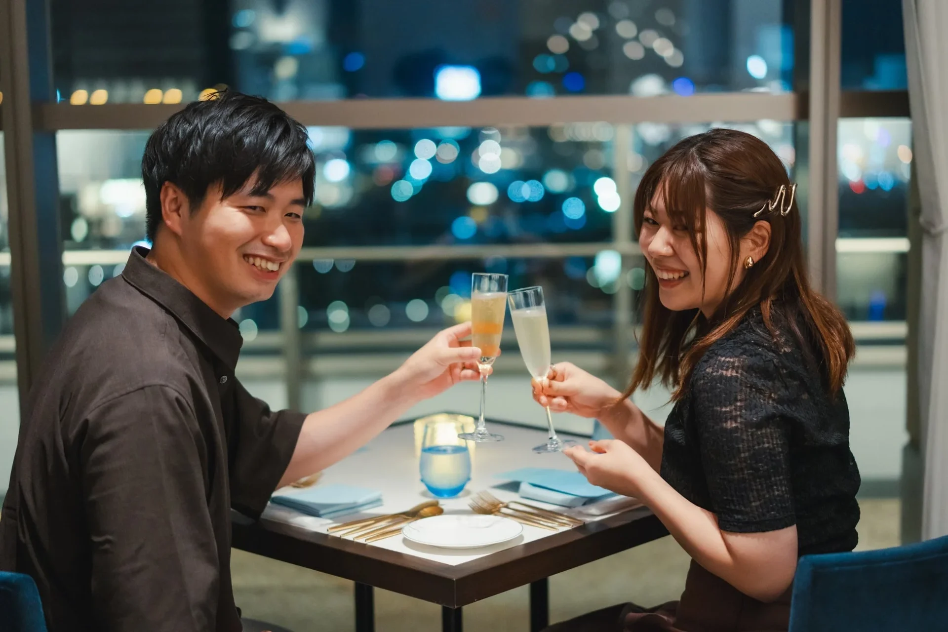 夜景が見えるレストランで、笑顔のカップルがシャンパンを片手に乾杯している瞬間です。二人の幸せそうな雰囲気が伝わる、ロマンチックなディナーシーンです。