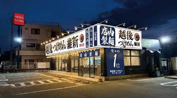 夜のラーメン店「越後つけめん 維新」の外観を捉えた画像です。店内製麺をアピールする看板や、「新潟つけめんランキング堂々の1位」と書かれた青い垂れ幕が目を引きます。