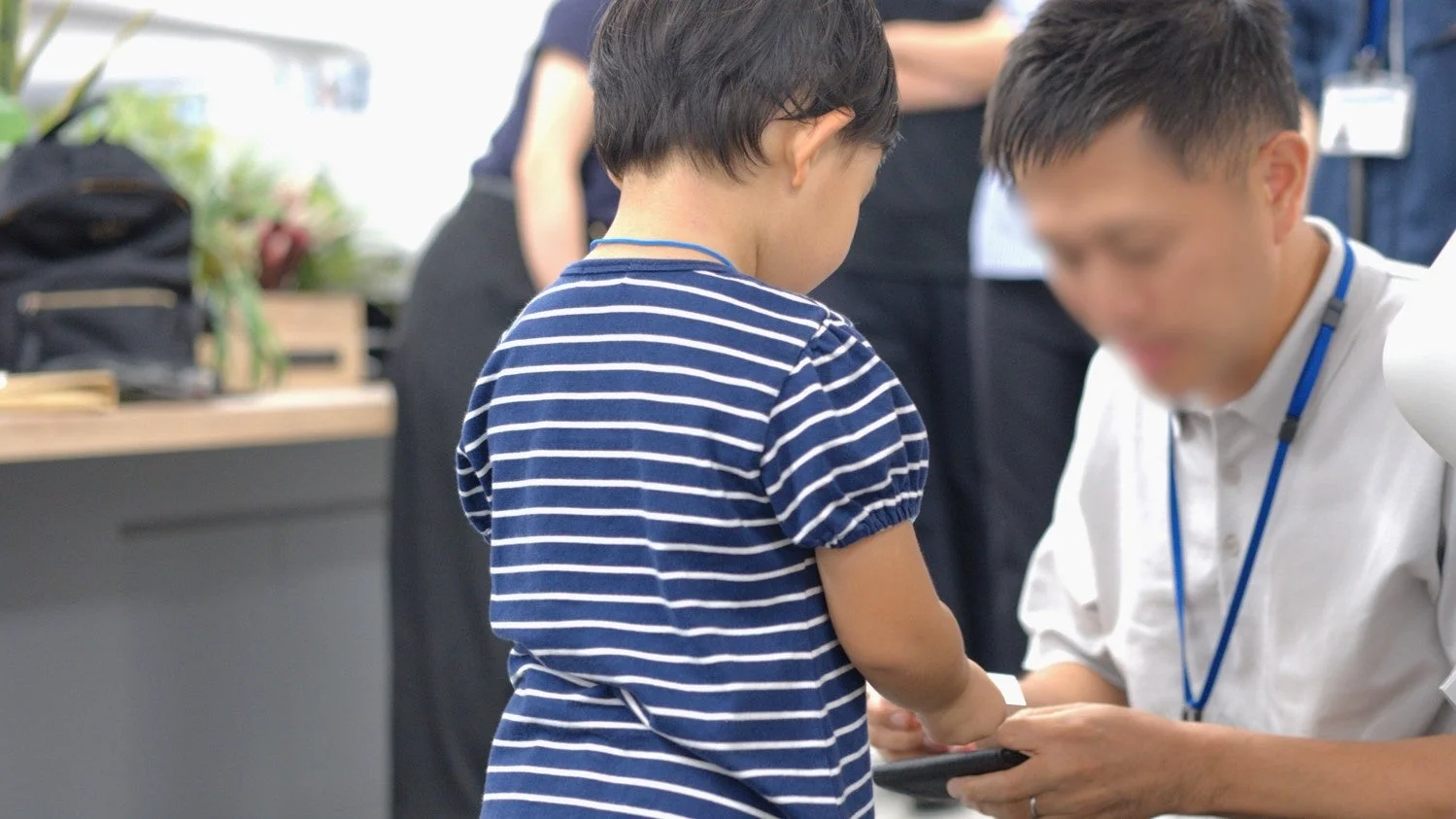 青と白のストライプのシャツを着た子供が、顔がぼかされた大人のそばに立っています。