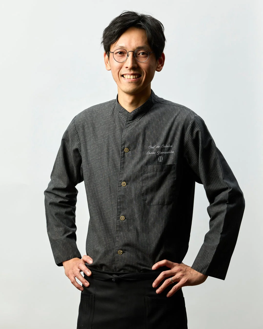 黒いコックコートを着用した日本人男性シェフが、白い背景の前で笑顔で立っているポートレート