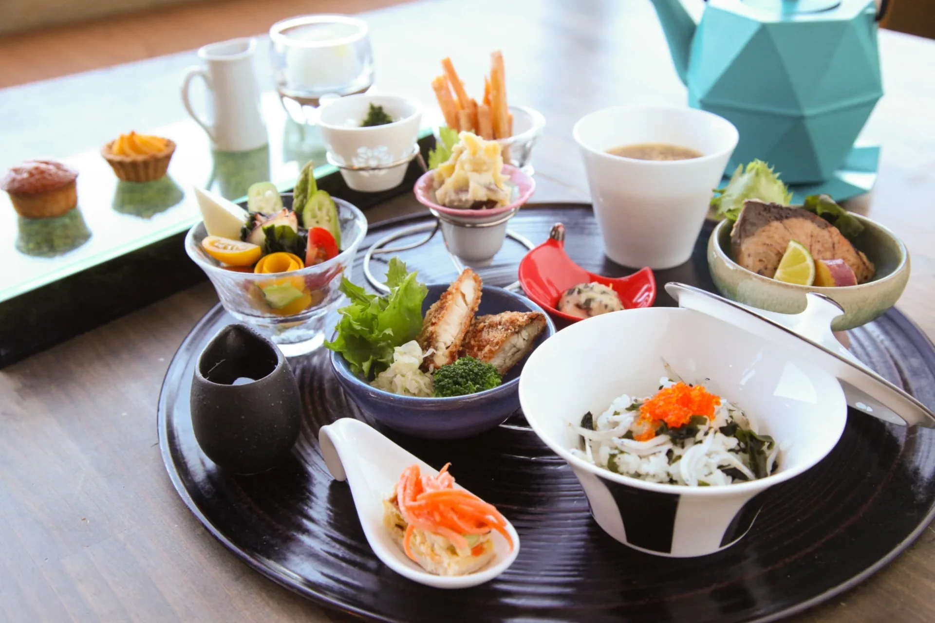 美しい器に盛られた彩り豊かな和食の定食。焼き魚、揚げ物、しらすといくらのご飯、サラダ、デザートなど品数豊富で、健康的な食事が楽しめます。モダンなデザインのティーポットも目を引きます。