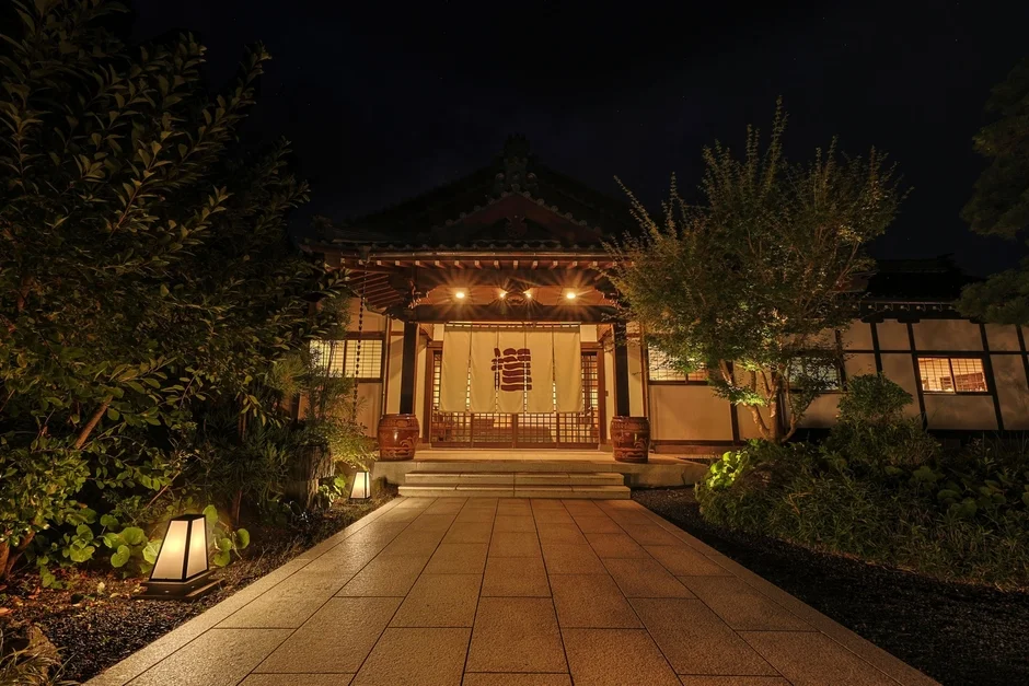 夜の帳が下りた日本の伝統的な建物が美しくライトアップされています。石畳の小道が入り口へと続き、左右には植栽と庭園灯が配置され、趣のある雰囲気を醸し出しています。暖簾には「福」の文字が見えます。