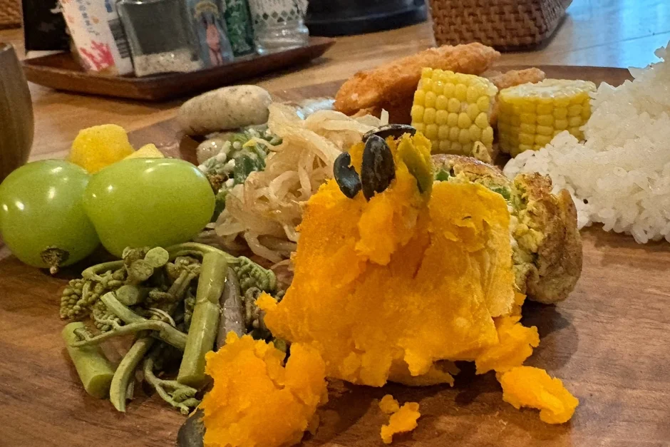木製の皿に盛り付けられた、かぼちゃペースト、ぶどう、とうもろこし、ご飯、様々な野菜が彩り豊かに並んだヘルシーなワンプレート料理。