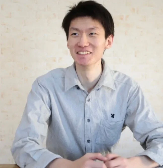 明るい色のシャツを着たアジア系の若い男性が、笑顔で横を向いている上半身のポートレート