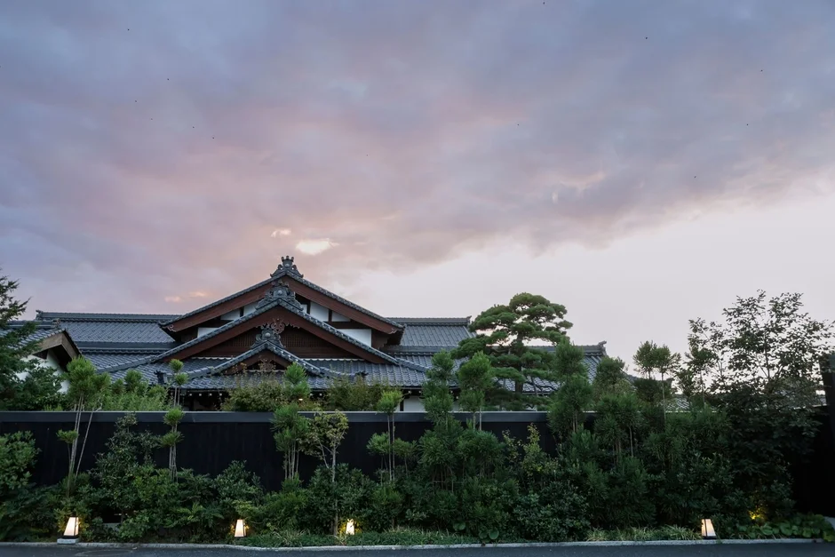 瓦屋根の伝統的な日本家屋が、手入れされた豊かな緑の庭園に囲まれています。夕暮れ時の柔らかな空が広がり、落ち着いた雰囲気です。足元には現代的な屋外照明が見えます。