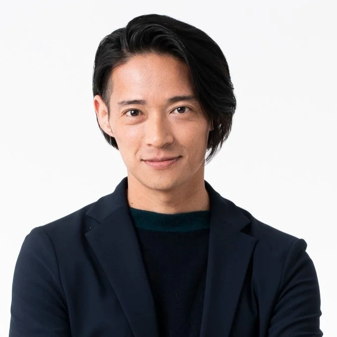 白い背景の前で、紺色のジャケットとセーターを着用した若いアジア人男性のポートレートです。彼はカメラに向かって穏やかに微笑んでおり、整えられた黒髪が特徴的です。