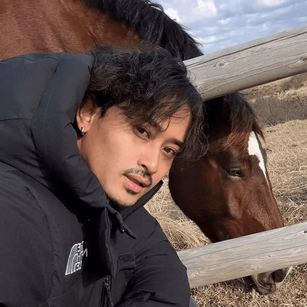 ダウンジャケットを着た男性が、茶色い馬と一緒にセルフィーを撮っています。馬は木製のフェンスの向こう側にいて、顔に白い模様が見えます。背景には乾いた草地が広がっています。