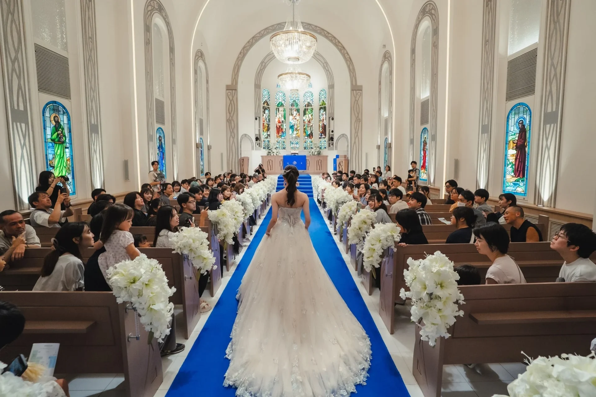 純白のウェディングドレスをまとった花嫁が、参列者に見守られながら美しいチャペルのブルーのバージンロードを歩いている結婚式の様子です。