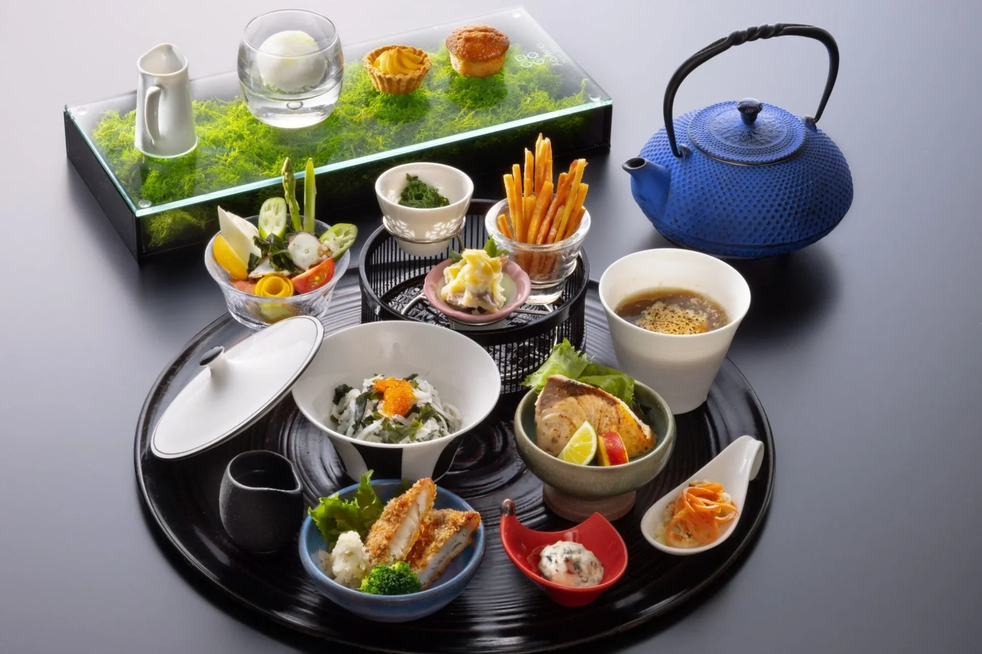 豪華で彩り豊かな和食のセットメニューが黒いお盆に美しく盛り付けられています。焼き魚、揚げ物、サラダ、汁物、デザートなど多様な料理が並び、青い鉄瓶も添えられた上品な食卓の様子です。