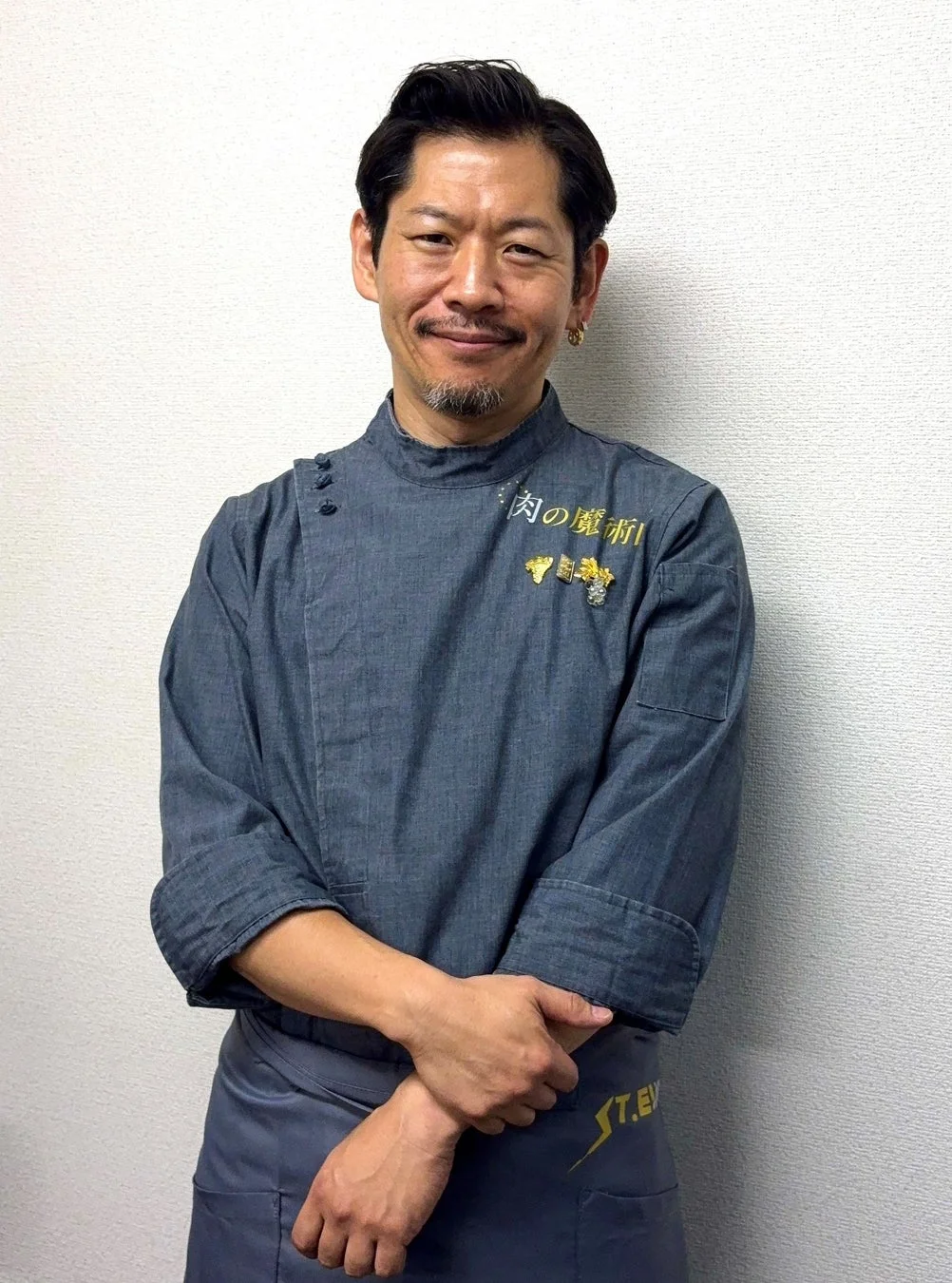 デニム風のシェフコートとエプロンを着用し、笑顔でカメラを見つめる日本人男性のポートレート