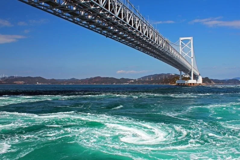 青い空の下、大鳴門橋の下に広がる鳴門の渦潮の壮大な景観。エメラルドグリーンの海面に力強い渦が巻いています。
