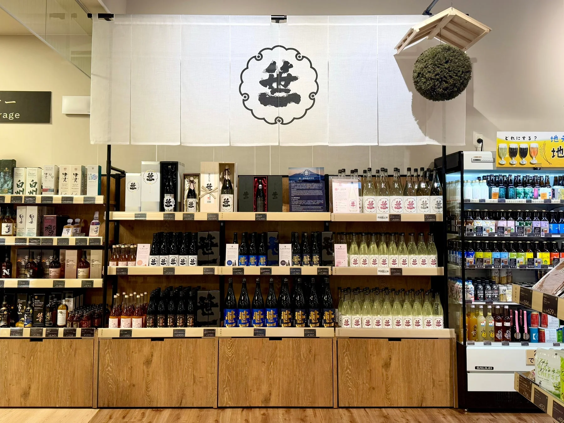 「豊」の文字が特徴的な日本酒が並ぶ酒屋の棚。上部には「豊」の暖簾が飾られ、様々な価格帯の日本酒やビール、その他の飲料が陳列されている。和風で豊富な品揃えの店内風景。