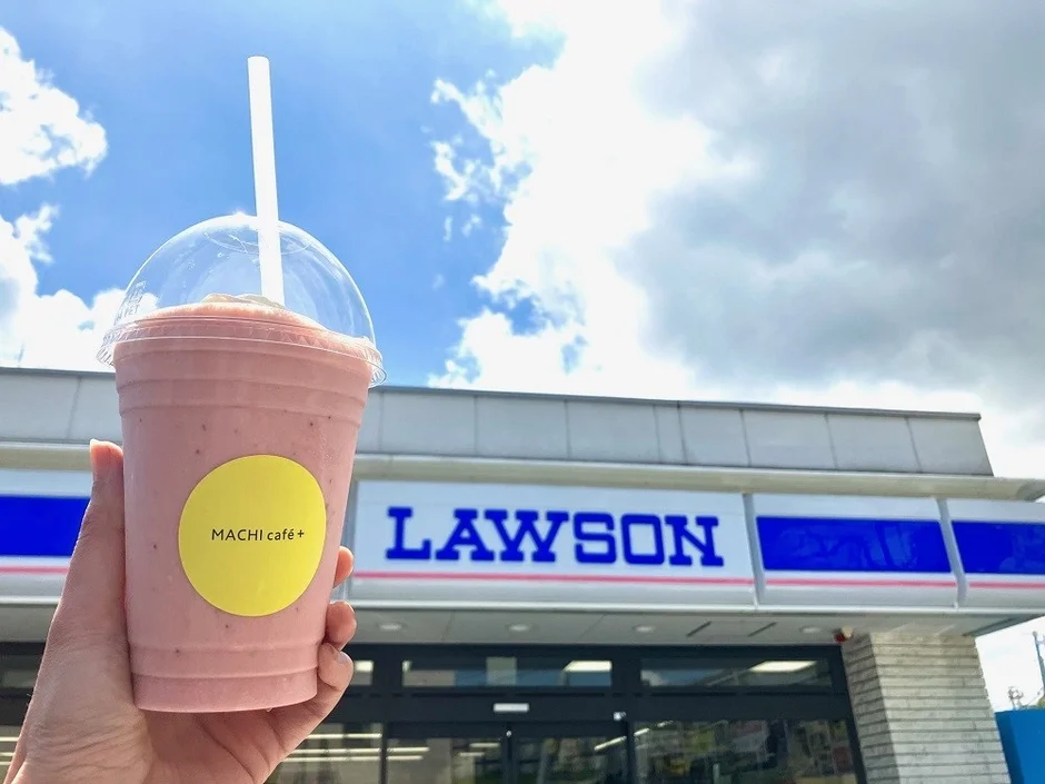 青空の下、ローソンの店舗を背景に、MACHI café+のピンク色のスムージーを手に持っている様子。夏にぴったりの冷たいドリンクです。