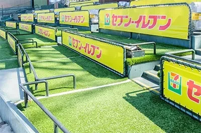 人工芝の通路と階段が特徴的なスタジアムの観客席エリアで、複数のセブン-イレブンの広告バナーが設置されている様子です。スポーツイベント会場の一角を示しています。