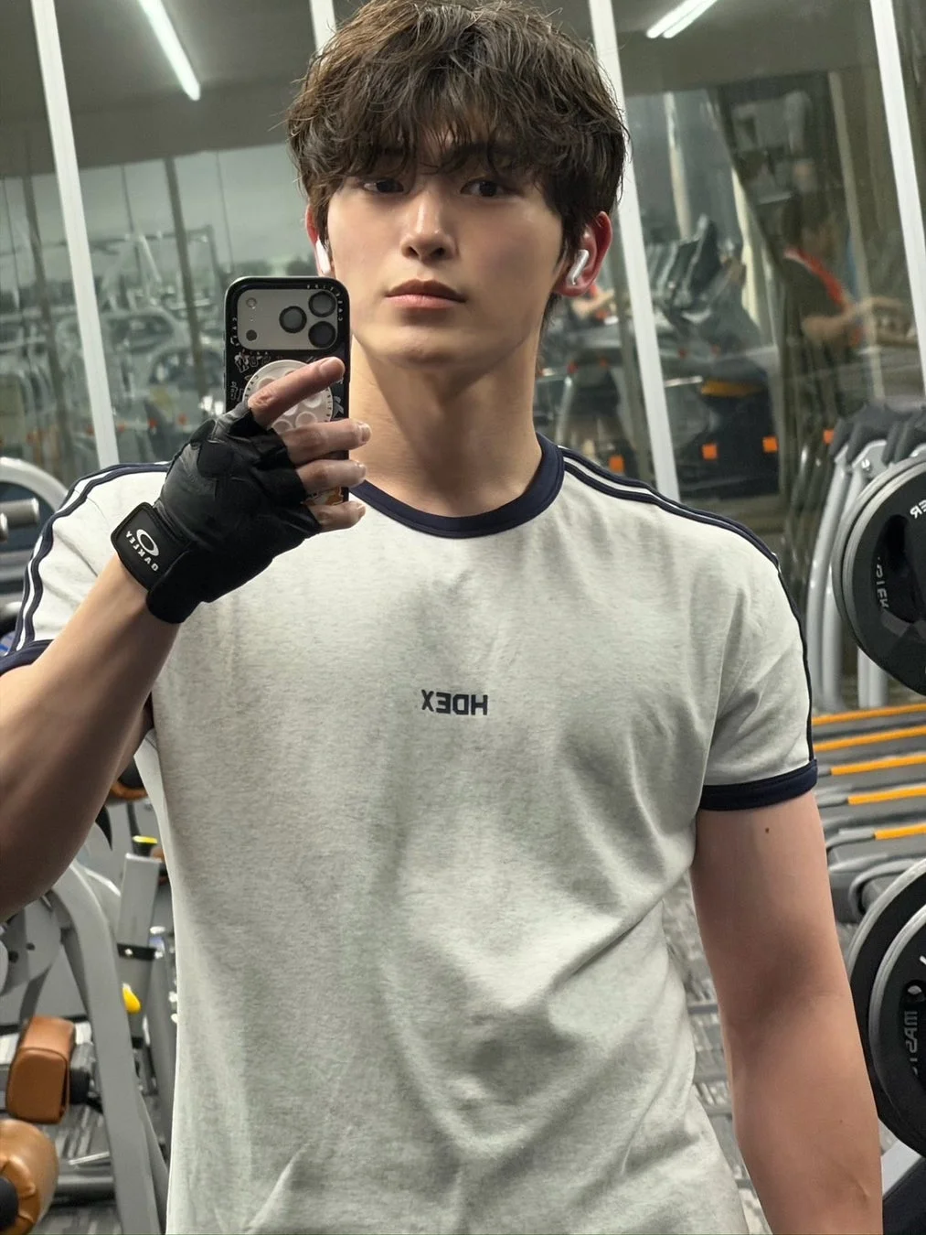 ジムでトレーニングウェアを着た若い男性が、鏡越しにスマートフォンで自撮りをしている様子。真剣な表情でカメラを見つめており、背景にはジムのマシンが見える。