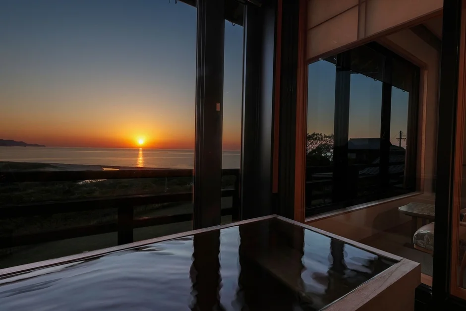 オーシャンビューの部屋から、海に沈む美しい夕日を望む光景です。手前には水が張られた木製の浴槽があり、夕焼け空が水面に映り込んでいます。贅沢な空間で、心安らぐひとときを過ごせるような雰囲気です。