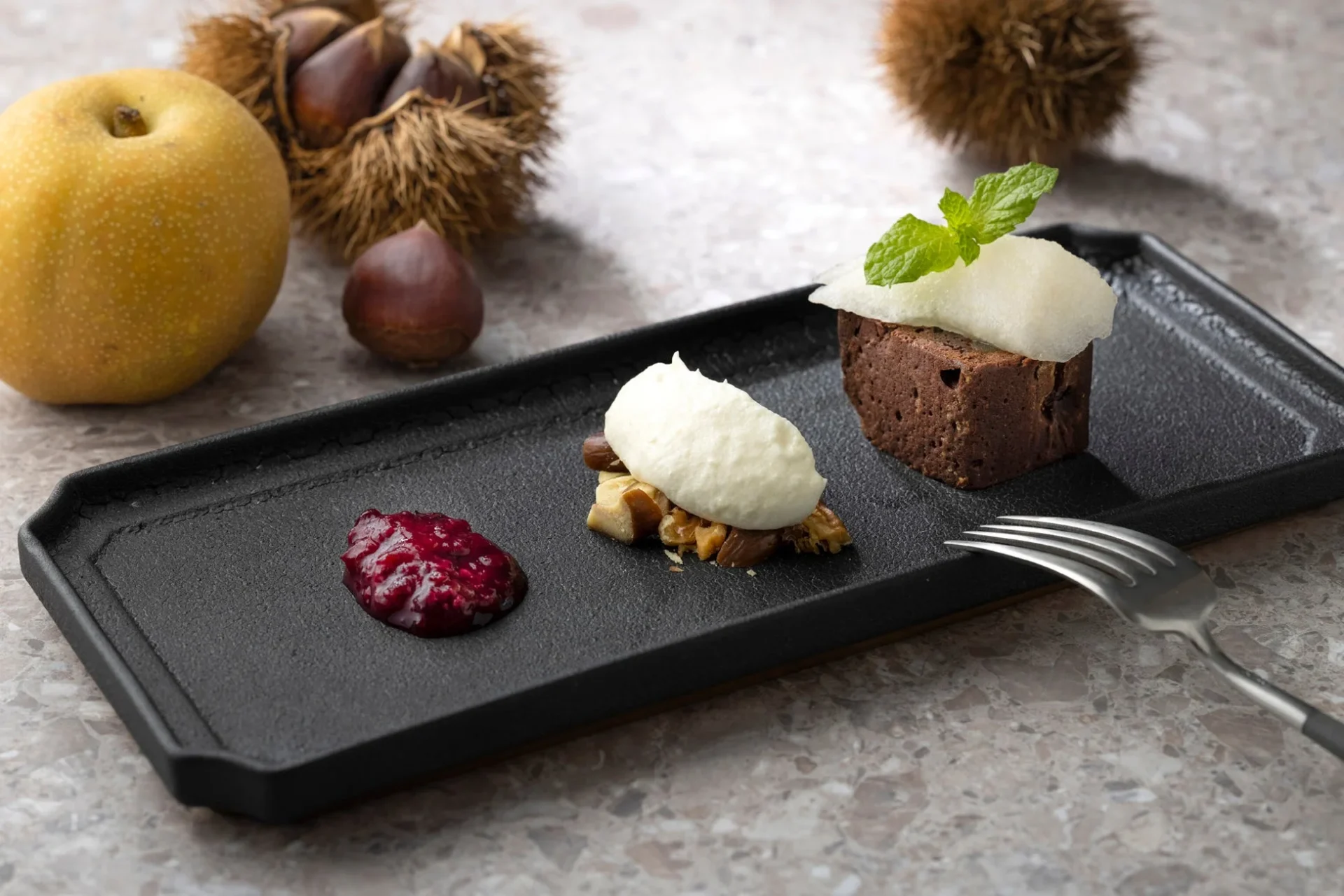 黒い皿に盛り付けられたチョコレートケーキとナッツ、ベリーソースのデザート。背景には洋梨と栗が置かれ、秋の味覚を表現した上品なスイーツの情景です。