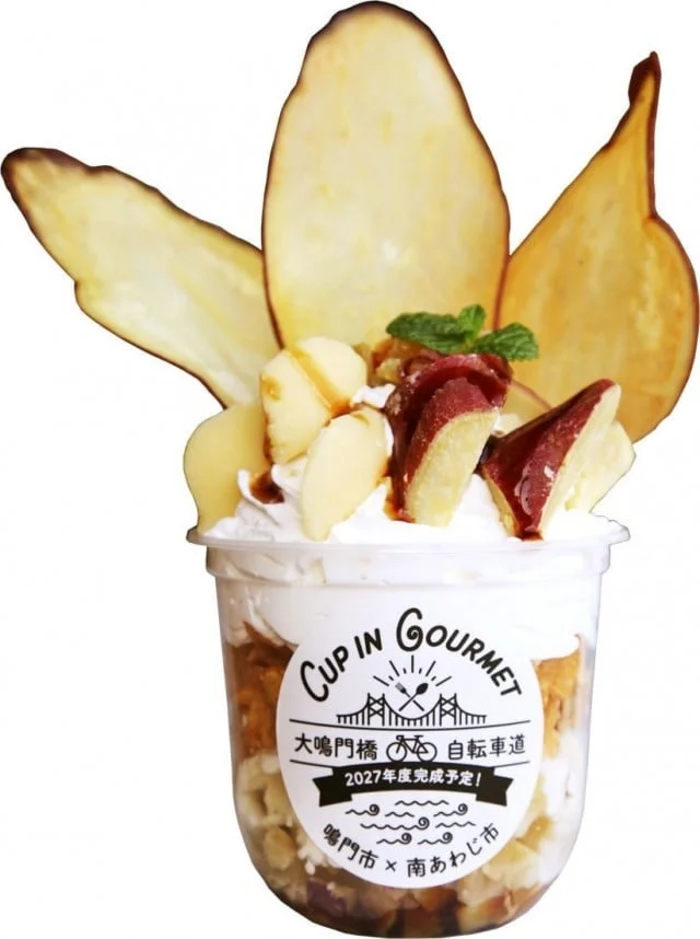 さつまいもをメインにしたカップ入りのスイーツです。ホイップクリームやナッツ、薄切りのさつまいもチップスが飾られています。カップのラベルには「CUP IN Gourmet」とあり、大鳴門橋自転車道の2027年度完成予定を告知しており、鳴門市と南あわじ市の地域プロモーションを兼ねた商品のようです。