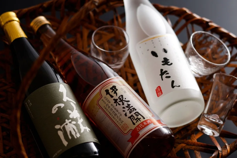 籠に並べられた3本の日本酒（伊根満開、人よろこびの酒、もとん）と数個の酒器が写っています。伝統的な和の雰囲気で、日本酒を楽しむ様子が伝わります。