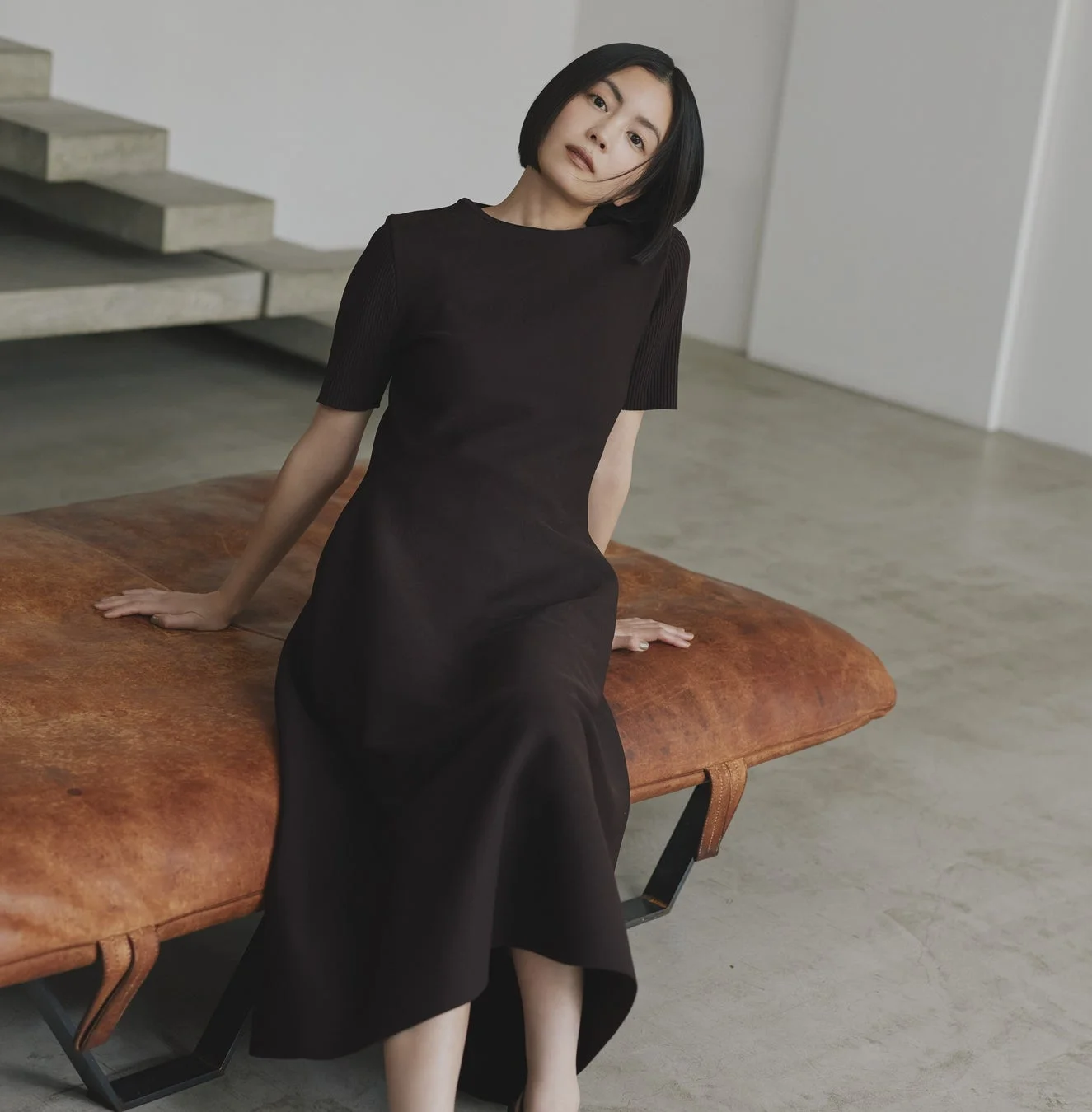 ダークブラウンのワンピースを着たショートヘアの女性が、革製のベンチに座っているポートレート
