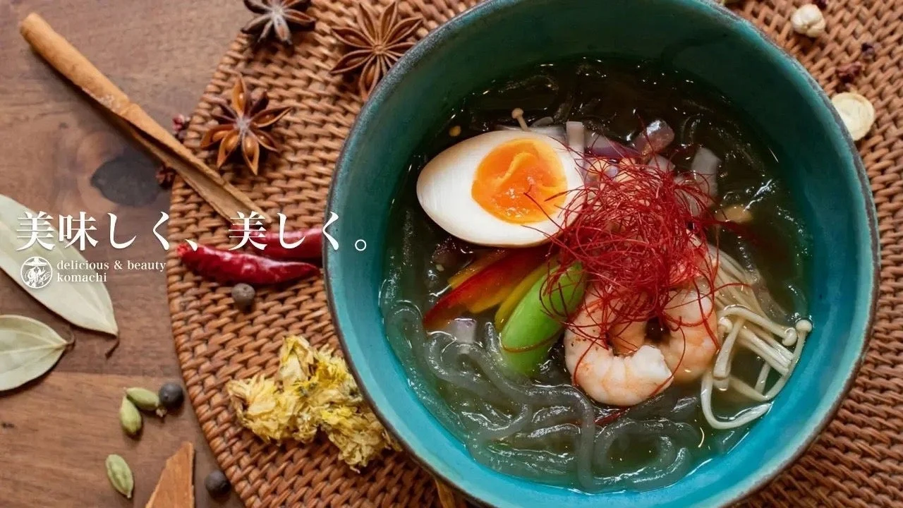 エビや半熟卵、キノコ、野菜、唐辛子の糸が彩り豊かに盛り付けられた麺料理のボウルが、様々な香辛料と共に編み目のマットの上に置かれている。美味しくて美しい、ヘルシーな一品を表現している。