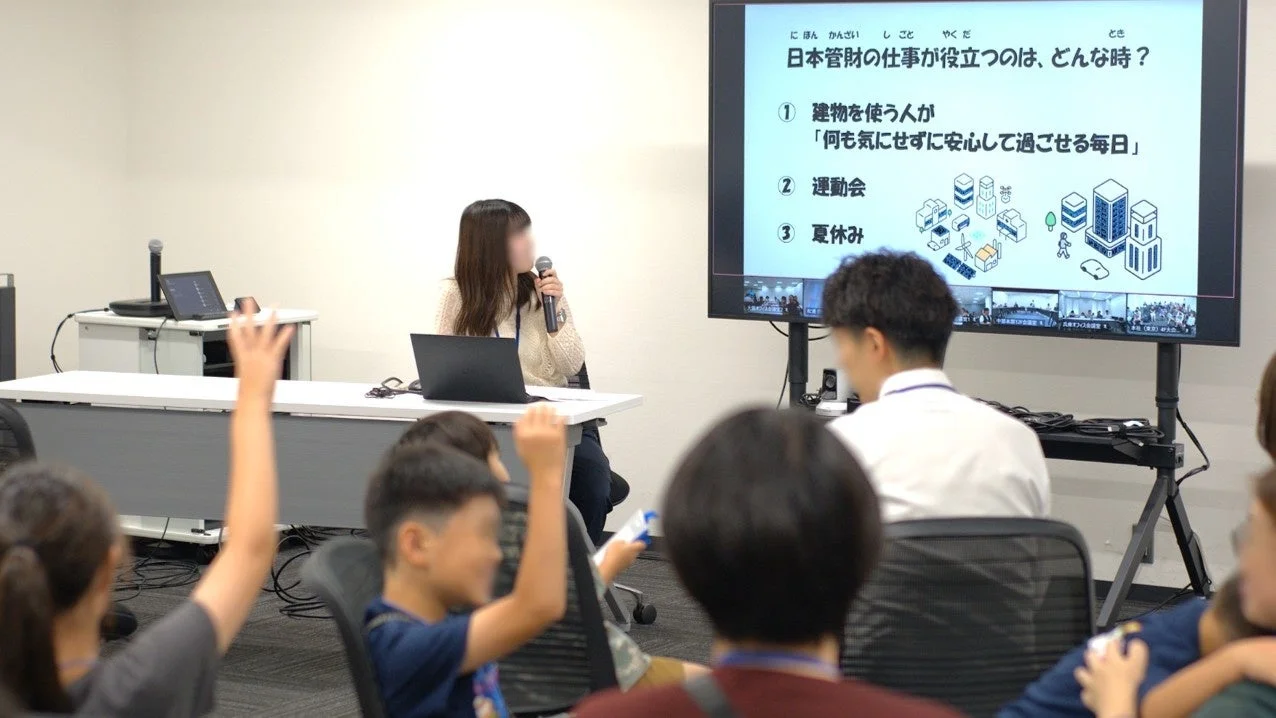 プレゼンテーションが行われている会場で、女性がマイクを持って説明しており、スクリーンには「日本管財の仕事が役立つのは、どんな時?」というテーマが表示されています。聴衆には子供たちが多く、数人が手を挙げています。