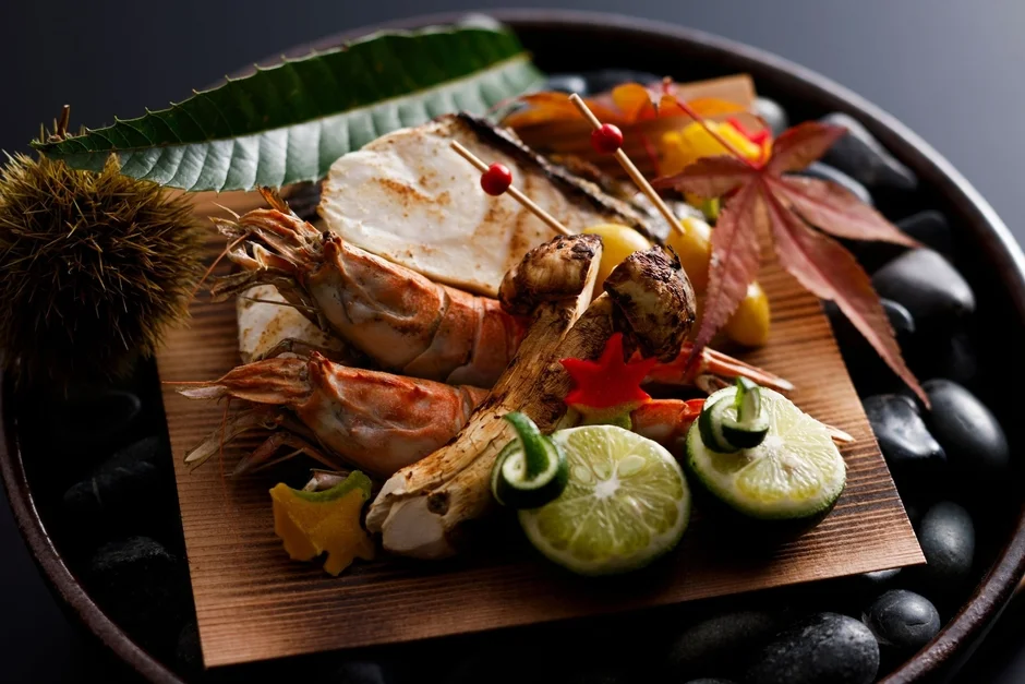 秋の味覚を盛り込んだ美しい日本料理。海老、魚、松茸のグリルを主役に、栗、すだち、紅葉が添えられ、季節感と上品さが際立つ一皿。黒い石で飾られた器が高級感を演出している。