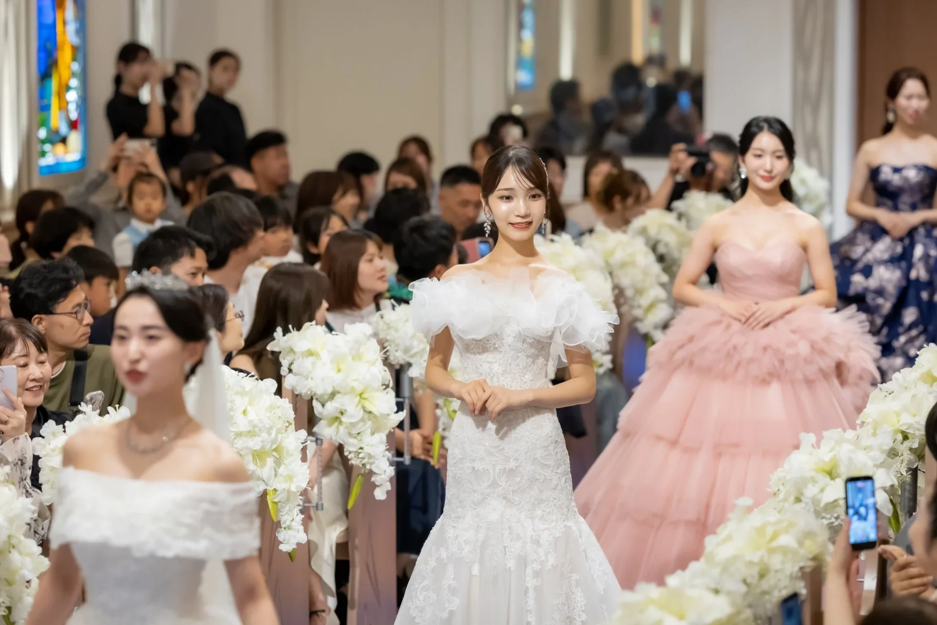 複数の女性がウェディングドレスやカラードレスを着用し、観客が見守る中でランウェイを歩いている様子です。ブライダルイベントやファッションショーの一場面と考えられます。