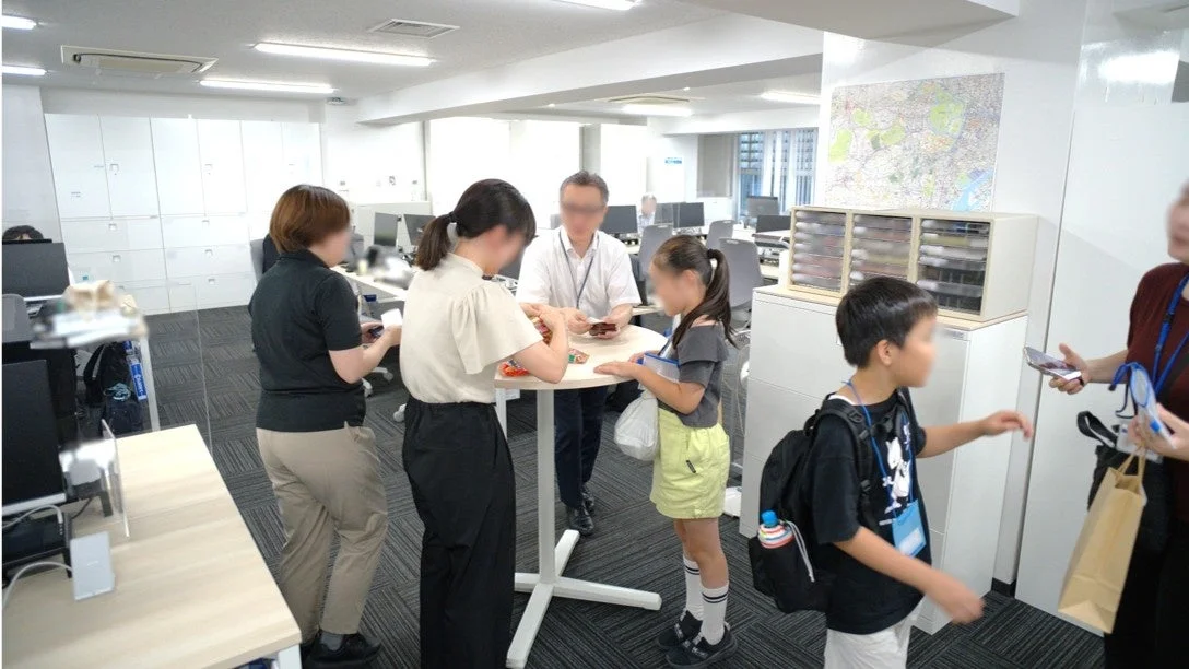 オフィス内で大人と子供たちが集まり、交流している様子です。丸いテーブルを囲んでお菓子のようなものをやり取りしており、職場見学やファミリーイベントの一環と見られます。