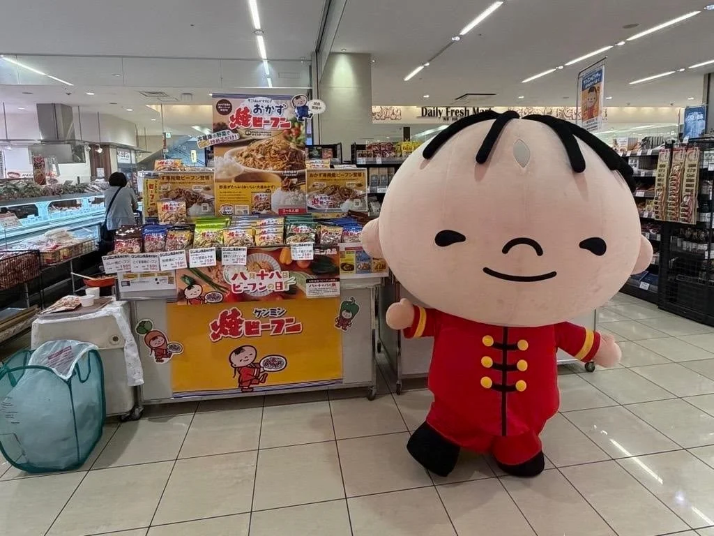 スーパーマーケットの売り場で、ケンミン焼ビーフンのマスコットキャラクターがプロモーションを行っている様子