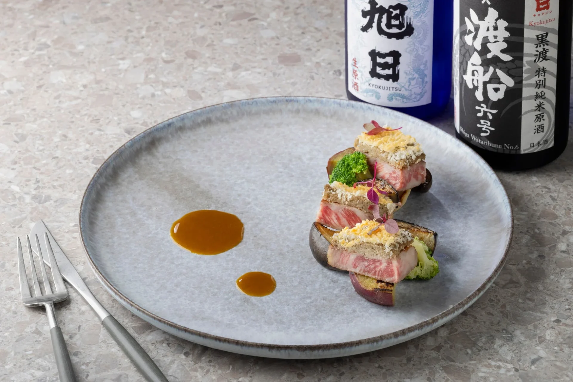 美しく盛り付けられた肉料理が写っており、奥には「旭日」と「渡舟」の日本酒ボトルが2本並んでいます。高級感のある食卓の様子です。