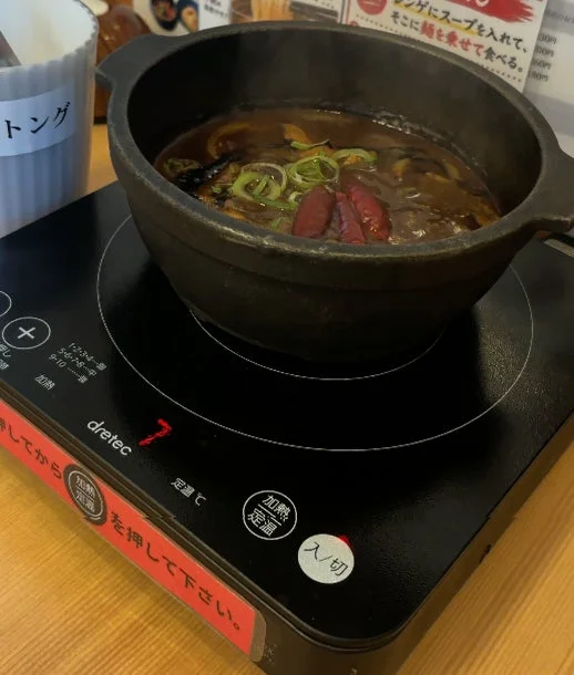 IHクッキングヒーターの上で、黒い鍋に入った熱々の煮込み料理が提供されている。ネギと唐辛子がトッピングされ、辛そうな中華風の料理に見える。飲食店での食事風景。