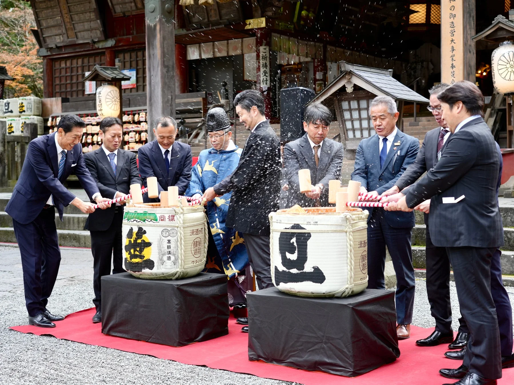 日本の伝統的な祝賀行事「鏡開き」の様子。複数の男性が酒樽を木槌で叩き割り、日本酒が飛び散っています。神社または寺院を背景に、お祝いムードが伝わる一枚です。