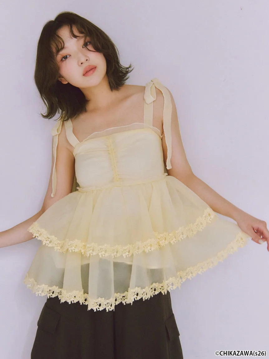 フリルとレースがあしらわれた淡いイエローのティアードトップスと黒いパンツを着用した女性モデル