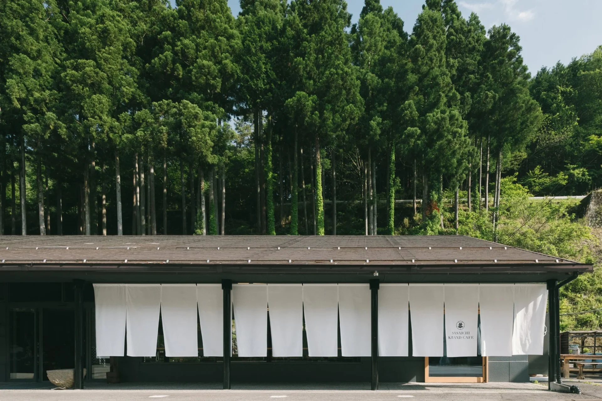 背の高い木々が茂る森を背景に建つ、白い暖簾がかかった和風の建物です。SASAICHI KIZND CAFEという文字が見え、自然豊かな場所にあるカフェを示唆しています。