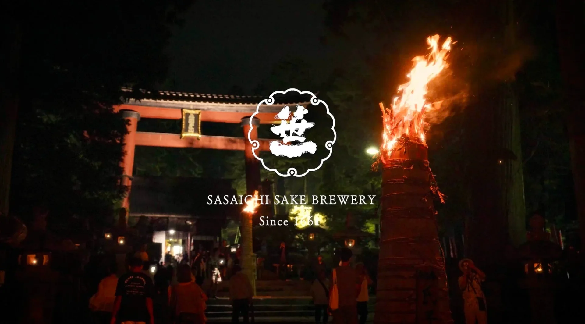 夜の神社で、大きな焚き火が燃え盛る中、人々が集まり祭りの雰囲気を楽しんでいます。鳥居と「笹一酒造」のロゴが見え、伝統的な日本のイベントの様子を捉えています。