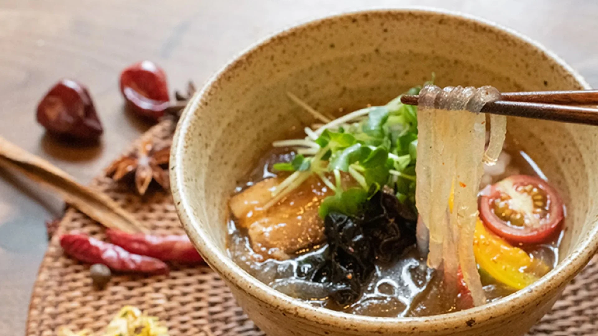 箸で持ち上げられた透明な麺が特徴的な、具だくさんの冷たい麺料理。トマト、海藻、スプラウトなどが彩り豊かに盛り付けられ、背景には香辛料が添えられています。健康的で食欲をそそる一品です。