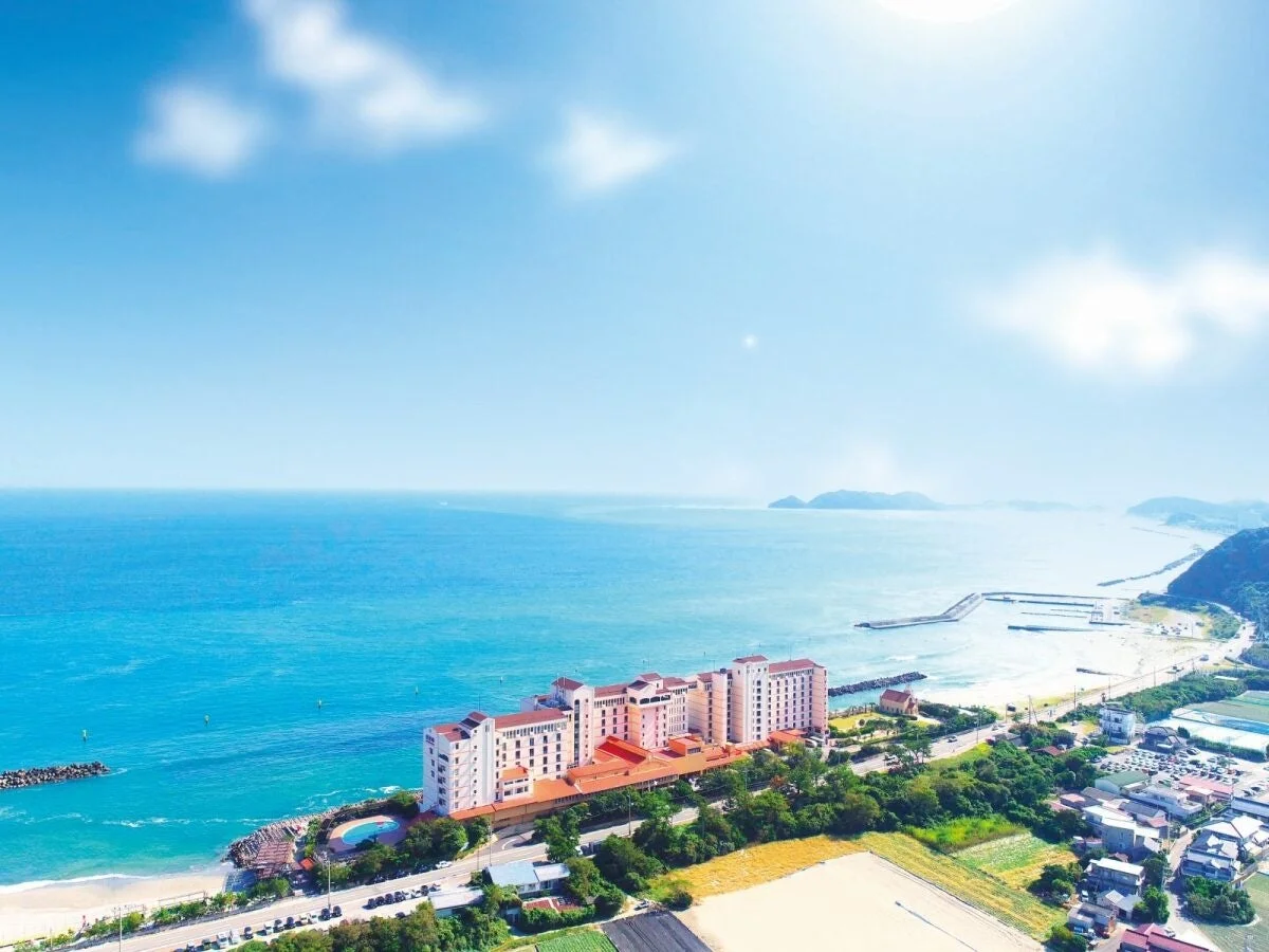 青い海と広がる海岸線に建つリゾートホテルを空撮した美しい景色。白い雲が浮かぶ青空の下、ビーチや周辺の街並みが広がり、穏やかな休日を思わせる。