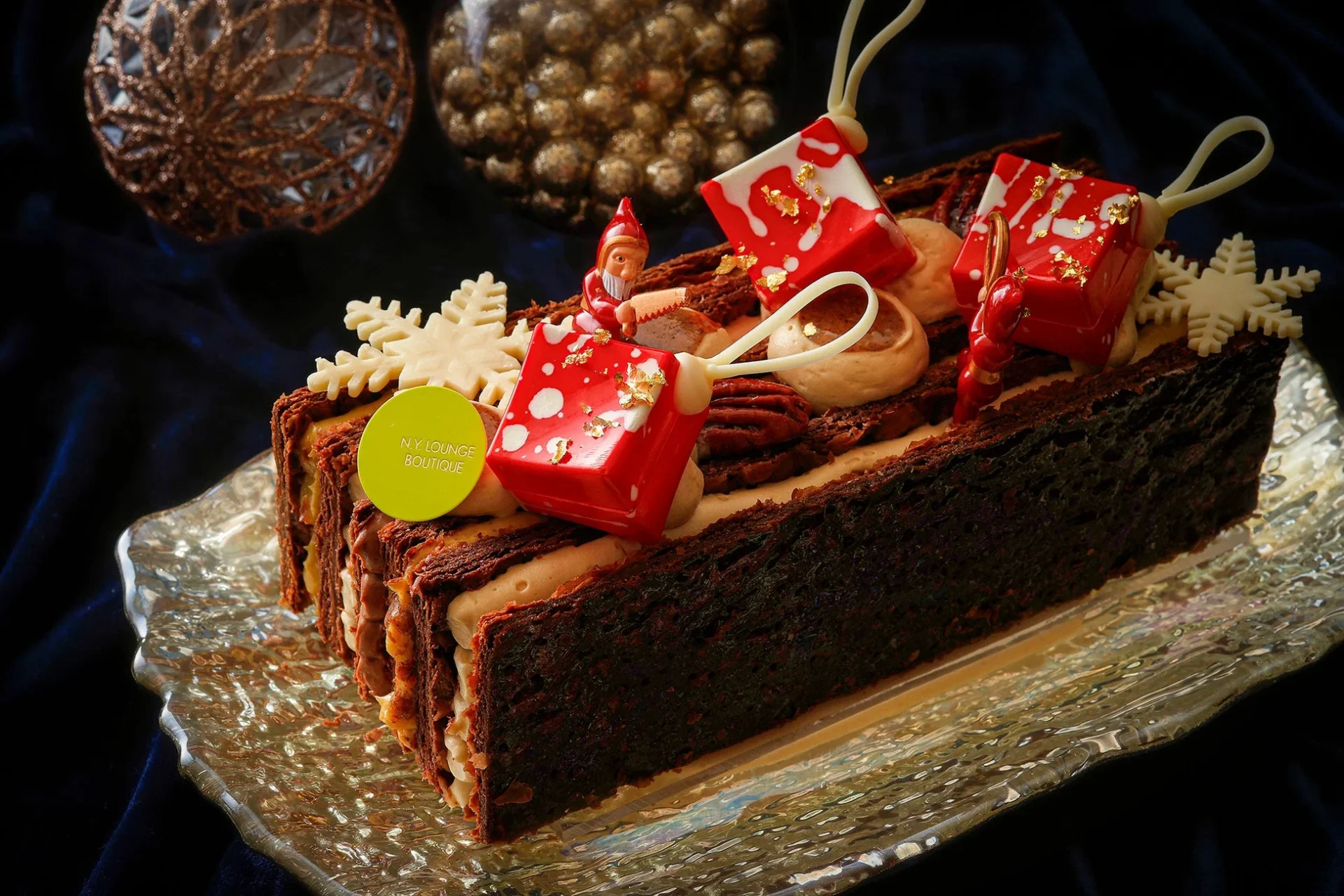 クリスマスをテーマにした豪華なブッシュドノエルです。チョコレートの生地にクリームやナッツが挟まれ、雪の結晶やサンタの飾り、赤いプレゼント型のチョコレートで華やかにデコレーションされています。
