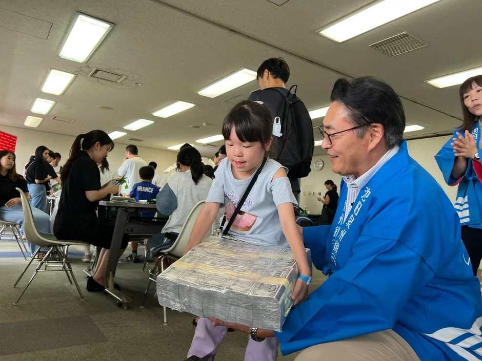 青い法被を着た男性が、大量の紙幣の束のようなものを運ぶ女の子を笑顔で手伝っている様子