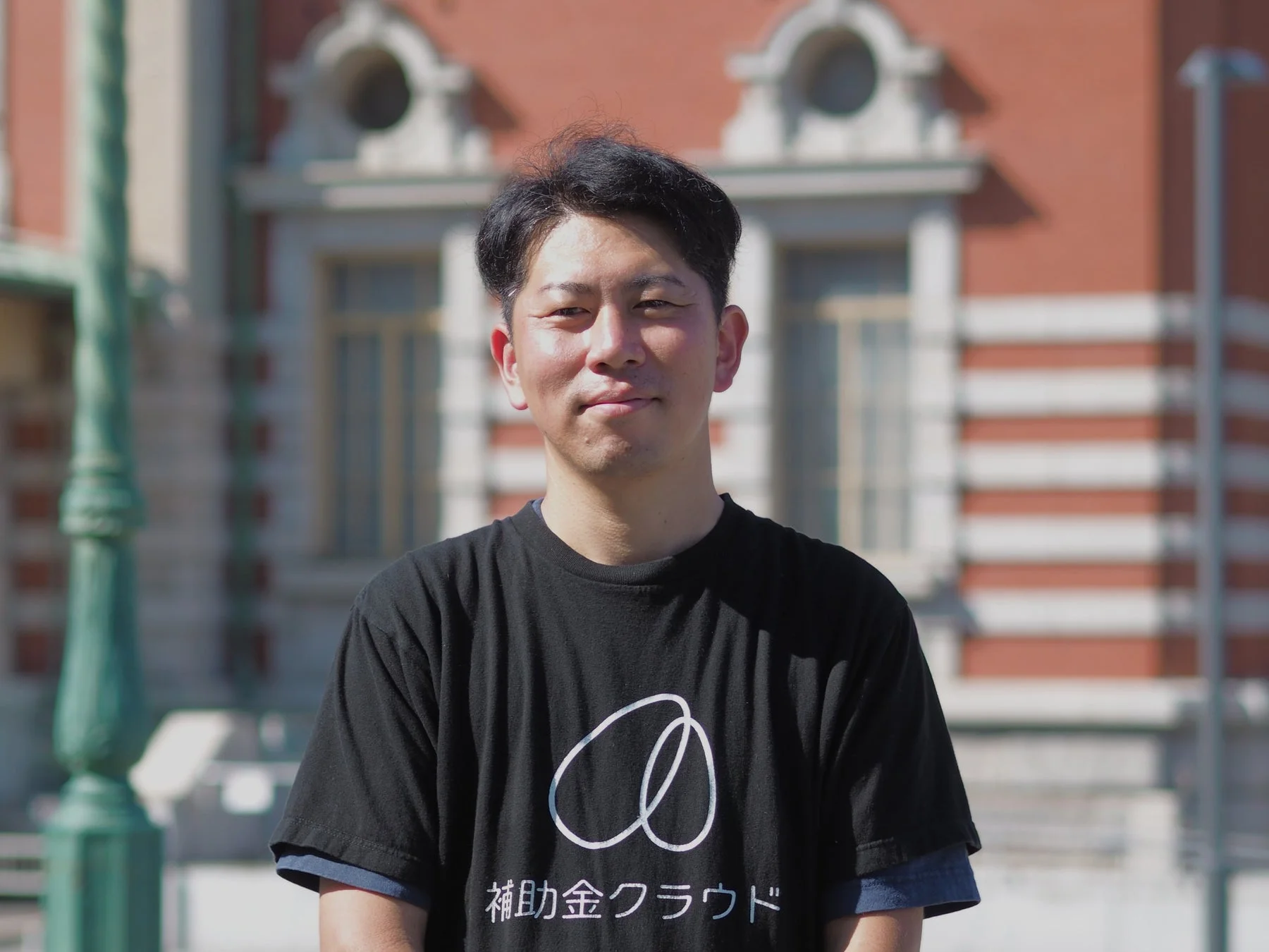 黒いTシャツを着たアジア人男性がカメラに笑顔を向けています。Tシャツには「補助金クラウド」とロゴがあり、背景には歴史的な赤レンガの建物が写っています。
