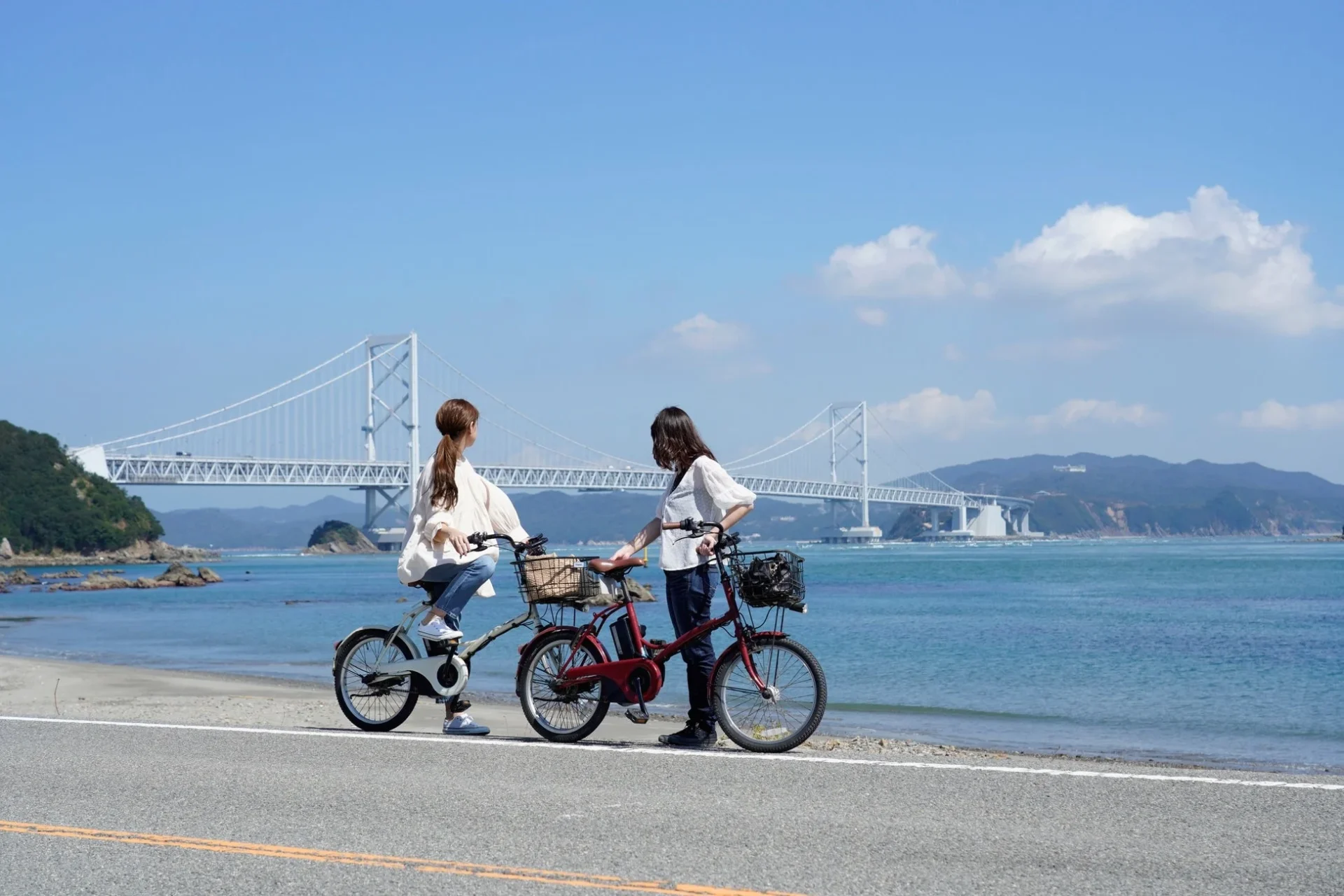青い空と海を背景に、自転車に乗った二人の女性が橋を眺めている風景です。しまなみ海道のような美しい場所でのサイクリングを楽しんでいる様子が伺えます。