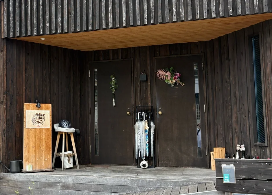 木製の外壁を持つ建物の玄関を捉えた画像。2つのダークブラウンのドアがあり、片方には花飾り、中央には傘立てが置かれている。