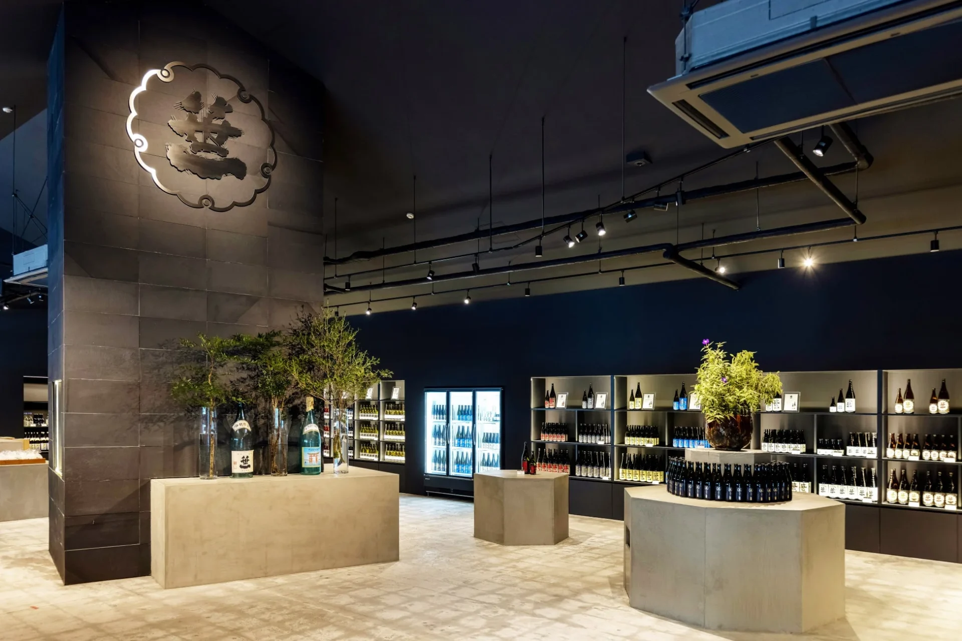 モダンで洗練された雰囲気の酒屋の店内。暗い色の壁とコンクリート製の什器が特徴で、棚には日本酒らしきボトルが多数並べられています。壁には光る和風のエンブレムがあり、店内には観葉植物が配置され、落ち着いた空間を演出しています。