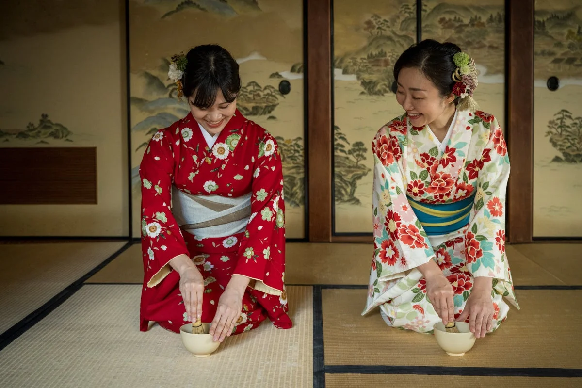 着物を着た二人の女性が、和室で笑顔で抹茶を点てている様子。日本の伝統的な茶道体験を楽しんでいる瞬間を捉えた一枚で、和やかな雰囲気が伝わります。