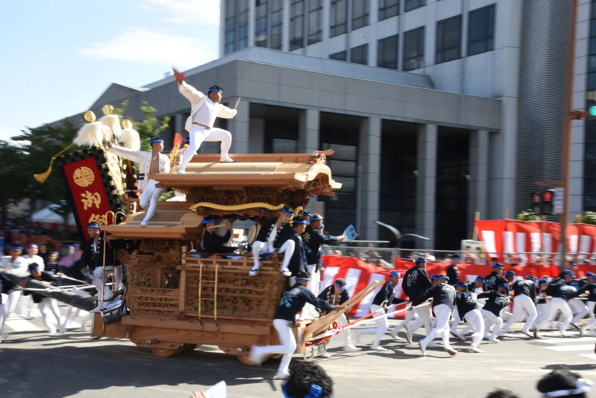 日本の伝統的な祭りで、多くの人々が装飾された木製の山車を引いている様子。山車の上では白い衣装の男性が扇子を持って舞い、周囲は活気に満ちている。