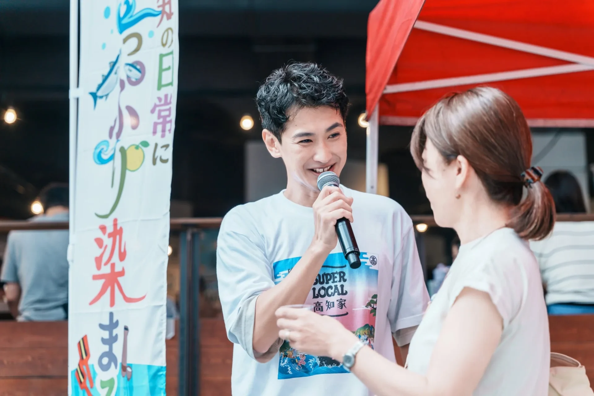 屋外イベントで男性がマイクを持ち、笑顔で女性と話す様子