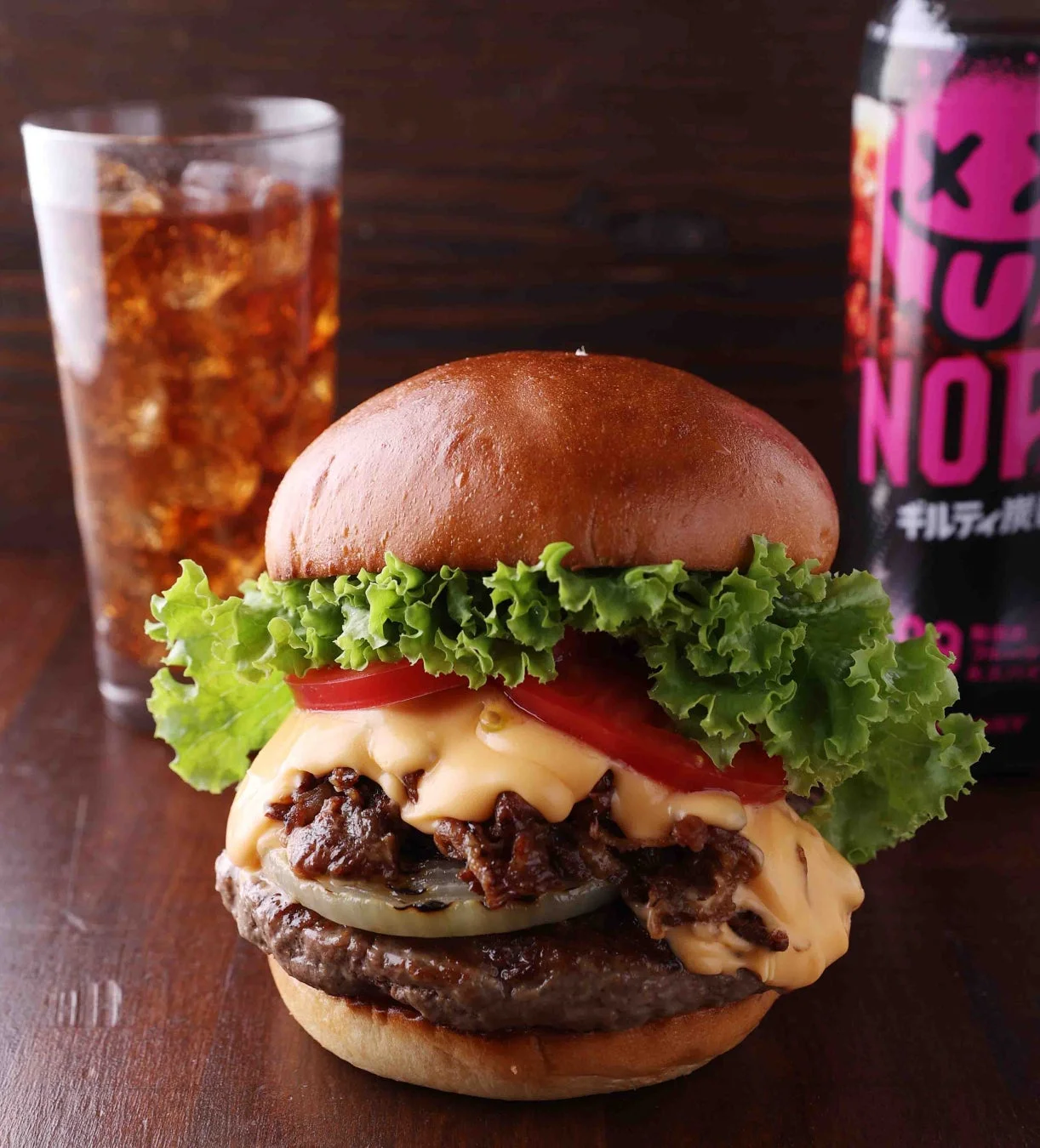 食欲をそそるチーズバーガーと、氷入りグラスのドリンク、そしてエナジードリンクの缶が写っています。バンズ、レタス、トマト、溶けたチーズ、肉厚なパティが特徴で、美味しそうな一食です。